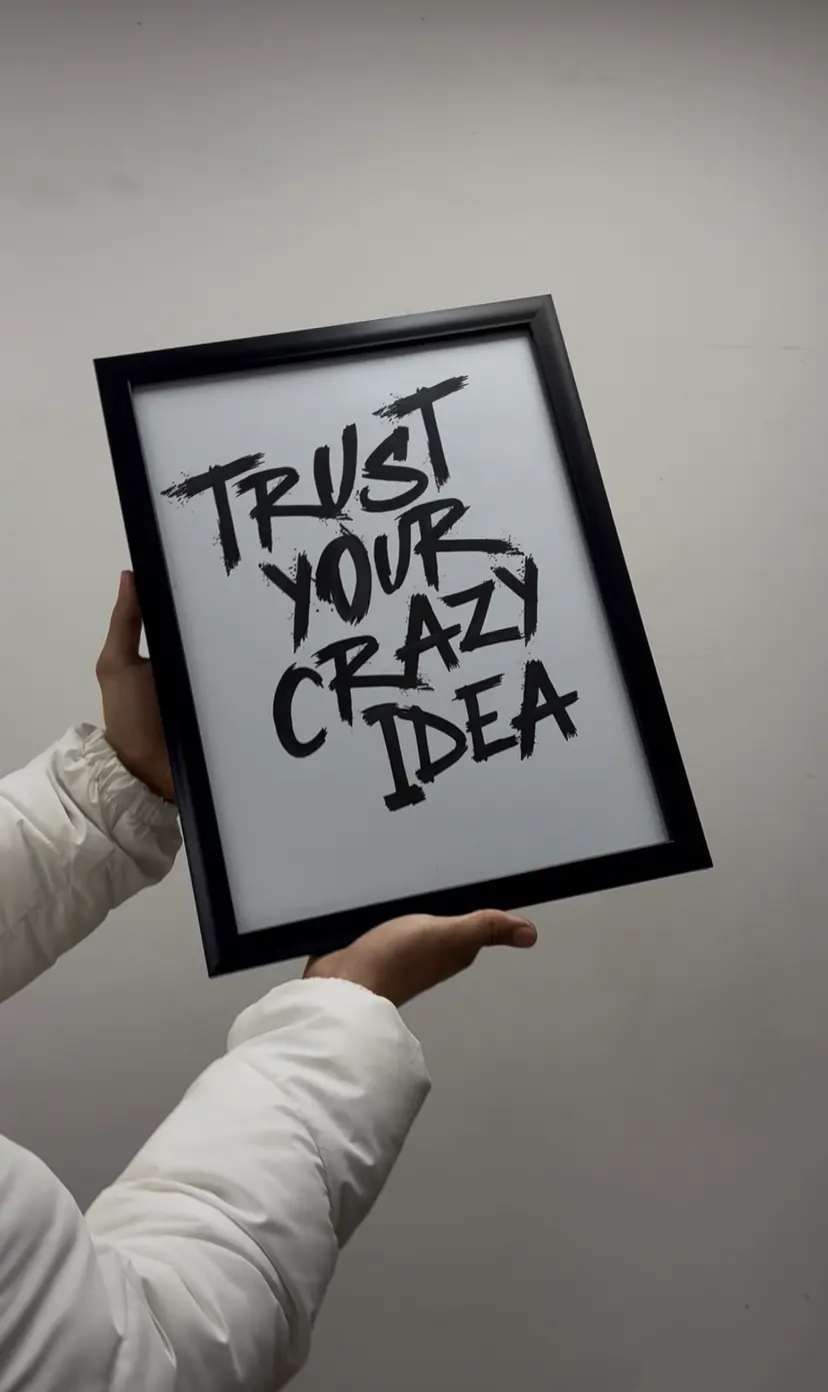 A black-framed Trust Your Crazy Idea poster held up in both hands